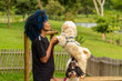 © Angela - Uma jovem negra de cabelos tingidos de azul, segurando as patas dianteiras de seu cachorro que está parado, em um parque bem arborizado, desfocado ao fundo.