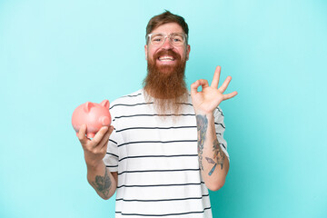 Wall Mural - Redhead man with long beard holding a piggybank isolated on blue background showing ok sign with fingers