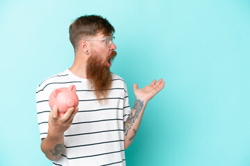 Wall Mural - Redhead man with long beard holding a piggybank isolated on blue background with surprise facial expression