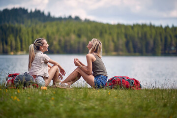 Wall Mural - Young female friends are sitting on the ground beside the lake while hiking the hills. Trip, nature, hiking