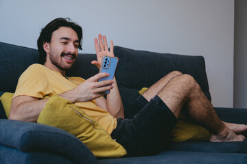 Young latin man on his living room couch making a video call on his cell phone. Copy space.