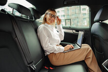 Attractive Businesswoman In Glasses Working On Laptop Sitting Car Backseat And Looking At Camera