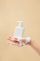 A female hand model holding an empty label pump bottle dispenser with white foam mousse. Beauty cleansing product mockup