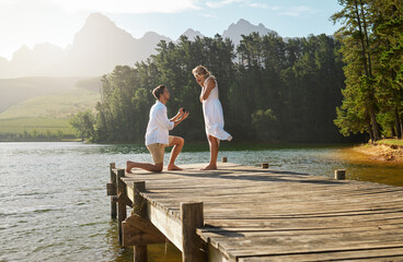 Canvas Print - Man, woman and engagement proposal by lake on vacation with surprise, wow or happiness in sunshine. Couple, marriage offer and ring in nature for romance, love and happy on holiday in summer by water