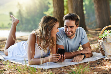 Wall Mural - Couple on picnic with smartphone, relax together in nature and social media with travel and bonding outdoor. Happiness, man and woman lying down and scroll internet in forest with smile