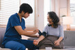 © kenchiro168 - A health visitor is measuring blood pressure a sick elderly woman who is sitting on a sofa at home. This scene illustrates the concept of home health care services.
