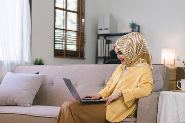 Wall Mural - Portrait of a beautiful Muslim millennial Asian businesswoman wearing a hijab working remotely from her home office while sitting on a sofa.