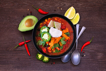 Chicken tortilla soup with tomatoes and black beans. Mexican food dish. Overhead view with frame of ingredients on a dark wood background.