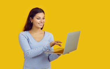 Happy beautiful young girl using notebook PC. Studio shot of pretty woman standing isolated on solid yellow background, holding her laptop computer, searching for something on the Internet and smiling