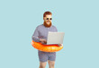 Funny man excited in anticipation of summer vacation using laptop booking tour on light blue background. Bearded chubby guy in striped leotard and with inflatable circle at waist is typing on laptop.