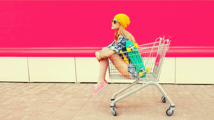 Poster - Stylish modern young woman sitting in trolley cart with skateboard looking away on colorful background, blank copy space for advertising text