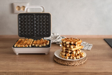 Cooking homemade waffles. A waffle iron with Belgian waffles and a stack of homemade waffles on a wooden kitchen table. 