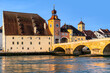 Danube embankment in Regensburg with the Old stone bridge and the Bridge clock tower in the early morning during golden hour, Bavaria, Germany