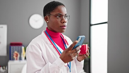 Wall Mural - African american woman doctor drinking coffee using smartphone at clinic