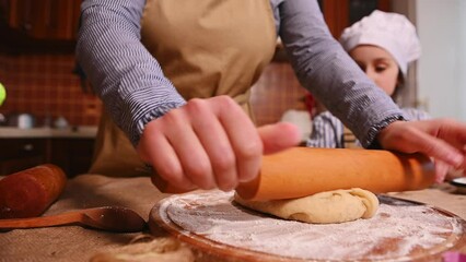 Wall Mural - Close-up of housewife woman in beige apron, rolls out dough with rolling pin. Her little daughter is at the table learning to cook. Mom and child cook gingerbread cookies together in the home kitchen