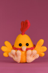 Wall Mural - Orange cockerel with bright red comb on head and beak crocheted, handmade art. Front view. Amigurumi chicken doll on pink background. Soft DIY toy made of cotton and natural wool.