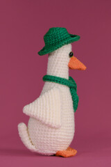 Wall Mural - Amigurumi duck doll on pink background. Soft DIY toy made of cotton. One white goose with orange beak wears green hat and scarf, crocheted from natural wool and cotton, handmade art. Side view.