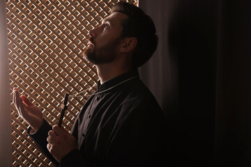 Wall Mural - Catholic priest in cassock holding cross and praying to God in confessional