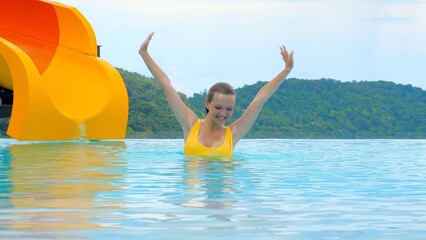 Smiling young woman in a yellow swimsuit dances in a pool on a hot sunny day with a water slide on the background. Female traveler having a good time and enjoying summer vacation.