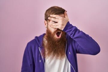 Wall Mural - Caucasian man with long beard standing over pink background peeking in shock covering face and eyes with hand, looking through fingers with embarrassed expression.