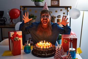 Wall Mural - Caucasian man with long beard celebrating birthday holding big chocolate cake celebrating victory with happy smile and winner expression with raised hands