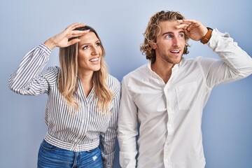 Wall Mural - Young couple standing over blue background very happy and smiling looking far away with hand over head. searching concept.