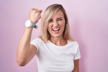 Poster - Young blonde woman standing over pink background angry and mad raising fist frustrated and furious while shouting with anger. rage and aggressive concept.