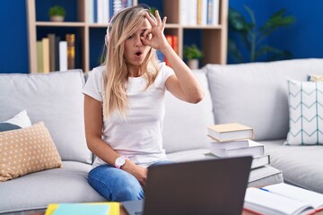 Poster - Young blonde woman studying using computer laptop at home doing ok gesture shocked with surprised face, eye looking through fingers. unbelieving expression.