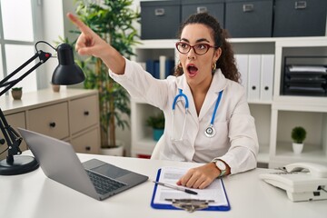 Canvas Print - Young hispanic woman wearing doctor uniform and stethoscope pointing with finger surprised ahead, open mouth amazed expression, something on the front