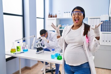 Poster - Young hispanic woman expecting a baby working at scientist laboratory celebrating mad and crazy for success with arms raised and closed eyes screaming excited. winner concept