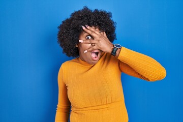 Sticker - Black woman with curly hair standing over blue background peeking in shock covering face and eyes with hand, looking through fingers with embarrassed expression.