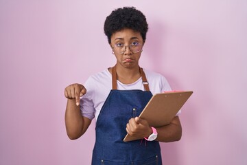 Poster - Young african american woman wearing professional waitress apron holding clipboard pointing down looking sad and upset, indicating direction with fingers, unhappy and depressed.