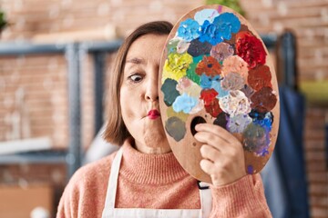 Poster - Middle age hispanic woman holding painter palette close to face making fish face with mouth and squinting eyes, crazy and comical.