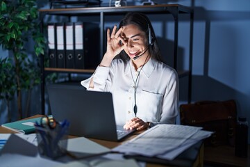 Poster - Young brunette woman wearing call center agent headset working late at night doing ok gesture with hand smiling, eye looking through fingers with happy face.