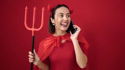 Wall Mural - Young beautiful hispanic woman wearing devil costume talking on smartphone over isolated red background