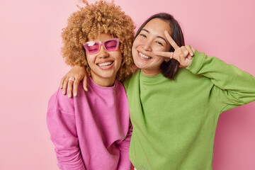 Wall Mural - Horizontal shot of positive mixed race young women friends smile and embrace make peace gesture have happy expressions dressed casually isolated over pink background. People and emotions concept