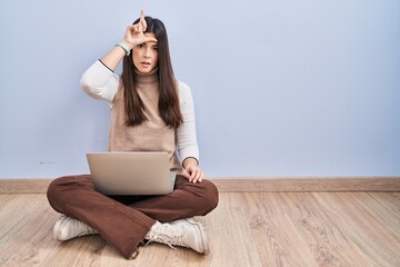 Canvas Print - Young brunette woman working using computer laptop sitting on the floor making fun of people with fingers on forehead doing loser gesture mocking and insulting.