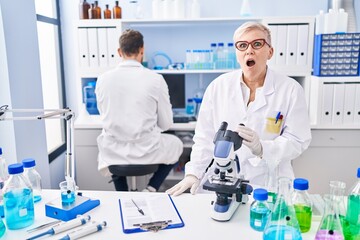 Wall Mural - Middle age woman working at scientist laboratory scared and amazed with open mouth for surprise, disbelief face