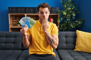 Poster - Young hispanic man holding chilean pesos covering mouth with hand, shocked and afraid for mistake. surprised expression