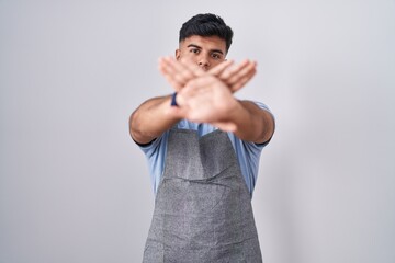 Poster - Hispanic young man wearing apron over white background rejection expression crossing arms and palms doing negative sign, angry face