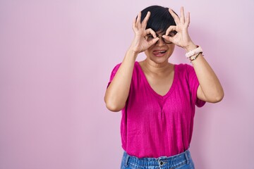 Sticker - Young asian woman with short hair standing over pink background doing ok gesture like binoculars sticking tongue out, eyes looking through fingers. crazy expression.