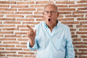 Canvas Print - Senior man with grey hair standing over bricks wall surprised pointing with finger to the side, open mouth amazed expression.