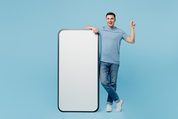 Wall Mural - Full body young happy man wear casual t-shirt big huge blank screen mobile cell phone with workspace copy space mockup area point index finger up isolated on plain pastel light blue cyan background.