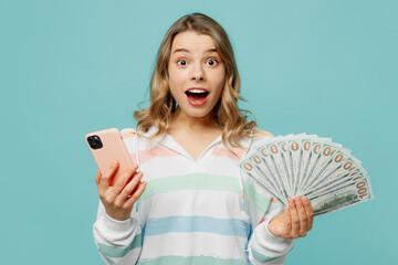 Wall Mural - Young amazed woman wear striped hoody use mobile cell phone hold in hand fan of cash money in dollar banknotes isolated on plain pastel light blue cyan background studio portrait. Lifestyle concept.