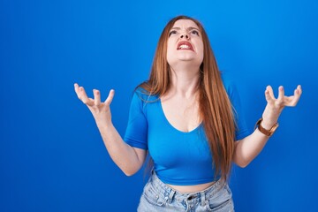 Sticker - Redhead woman standing over blue background crazy and mad shouting and yelling with aggressive expression and arms raised. frustration concept.