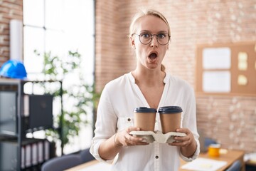 Canvas Print - Young caucasian woman working at the office holding coffee cups afraid and shocked with surprise and amazed expression, fear and excited face.