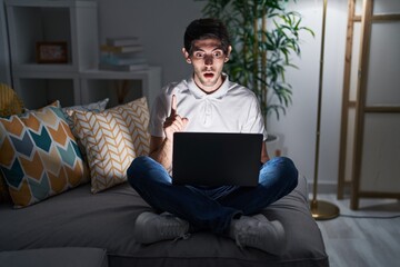 Poster - Young hispanic man using laptop at home at night amazed and surprised looking up and pointing with fingers and raised arms.