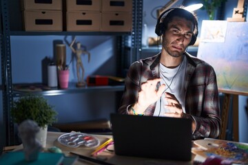 Poster - Young hispanic man sitting at art studio with laptop late at night disgusted expression, displeased and fearful doing disgust face because aversion reaction. with hands raised