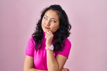 Sticker - Young asian woman standing over pink background looking confident at the camera smiling with crossed arms and hand raised on chin. thinking positive.