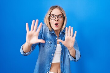 Poster - Young caucasian woman standing over blue background afraid and terrified with fear expression stop gesture with hands, shouting in shock. panic concept.